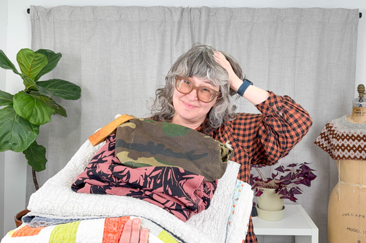 Jen Beeman in rust plaid Poppy Dress holding pile of mending projects with plants and dress form in background