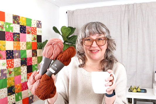 Jen Beeman in white handknit sweater holding pink and rust Isager Jensen yarn with mug, Heather Ross patchwork quilt behind