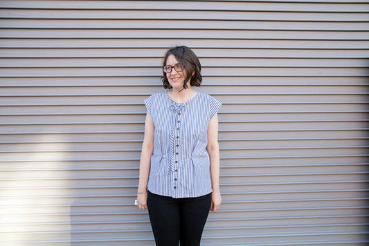 Person wearing a blue and white striped sleeveless top with lace-up neckline and black pants against a corrugated metal wall