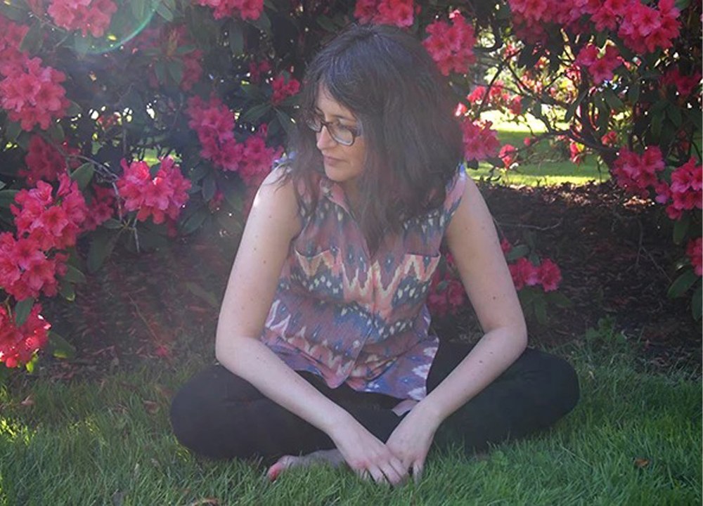 Woman sitting cross-legged on grass in a sleeveless ikat print Archer beside blooming pink rhododendron bushes