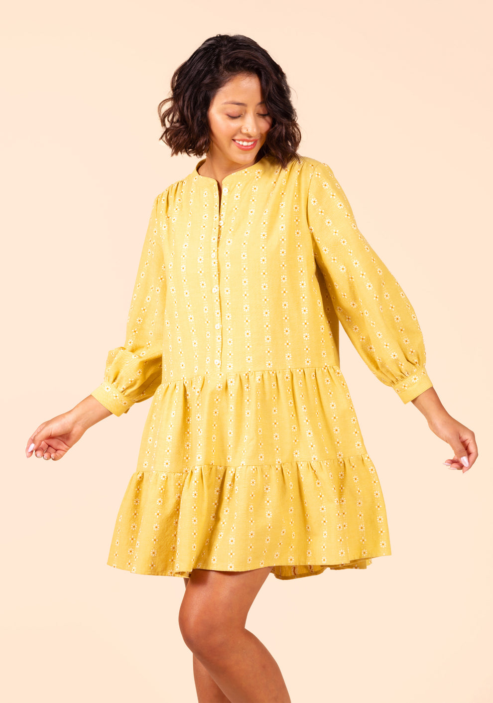 Model turning in Poppy Dress View B with tiered skirt and gathered sleeves in yellow daisy fabric.