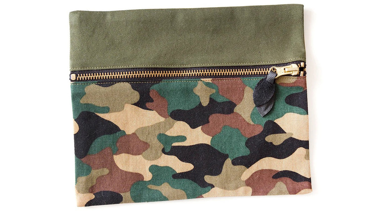 Completed travel pouch in camo and olive green fabrics with exposed brass zipper and black leather pull laid flat