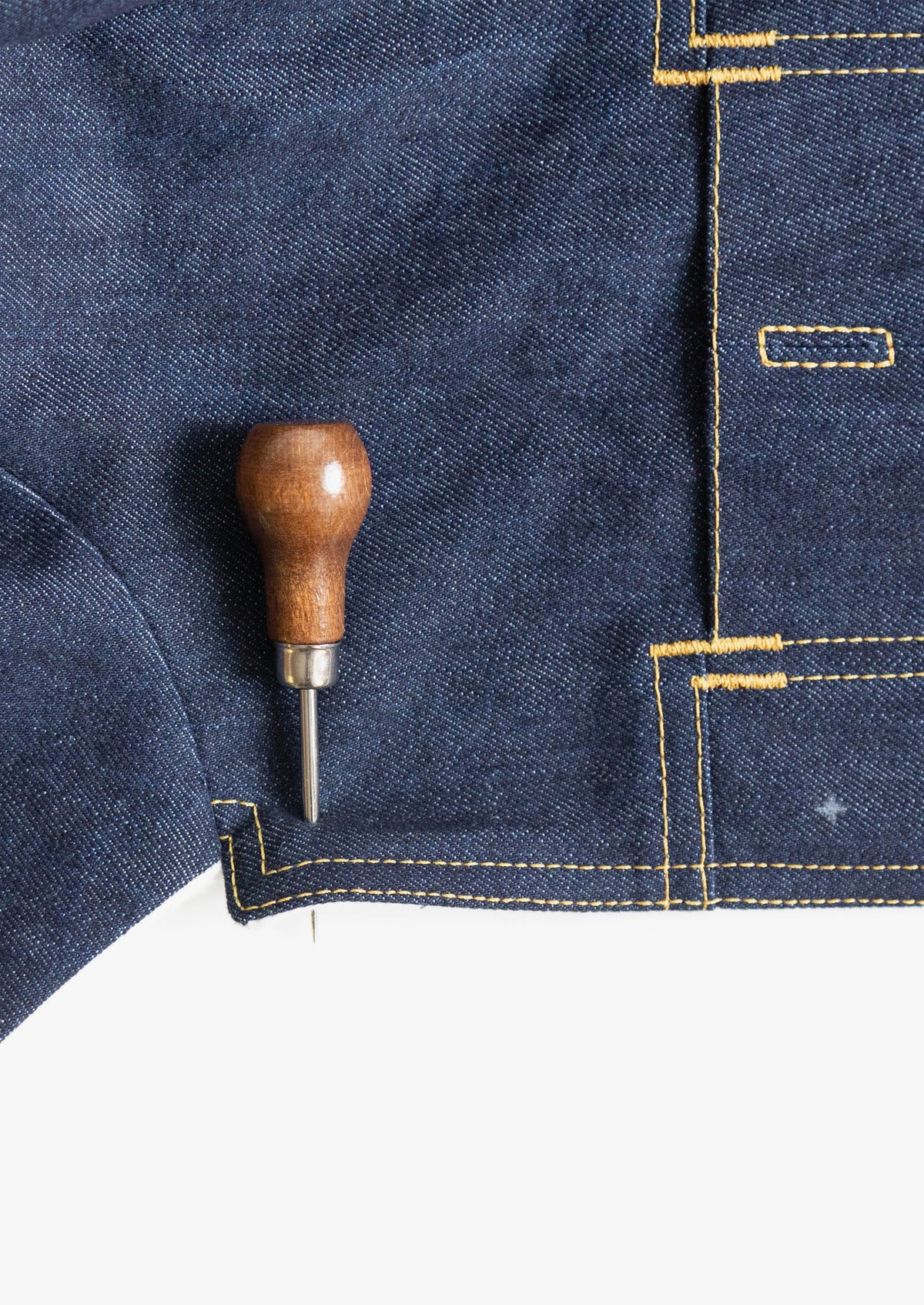 Awl marking position on dark denim fabric with visible yellow topstitching.