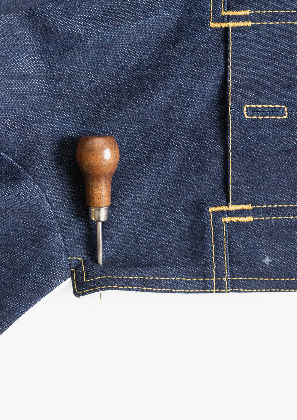 Awl marking position on dark denim fabric with visible yellow topstitching.